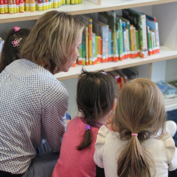 A woman sits in front of a bookshelf with three children and reads to them. The children listen attentively., &copy; Stadtbibliothek Stuttgart - Leseohren e.V.