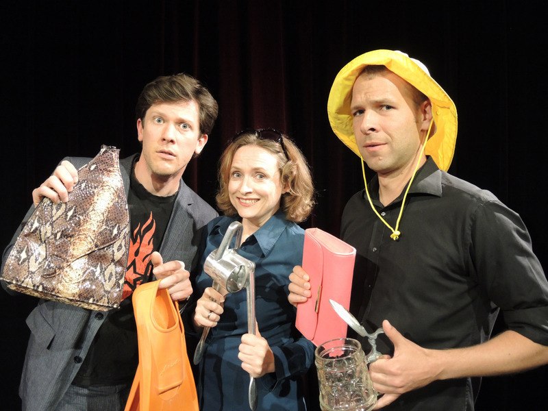 Three people stand on a stage holding bizarre objects such as a handbag, a lemon squeezer and a beer mug with cutlery., © Theaterhaus Stuttgart e.V. Three people stand on a stage holding bizarre objects such as a handbag, a lemon squeezer and a beer mug with cutlery., © Theaterhaus Stuttgart e.V.