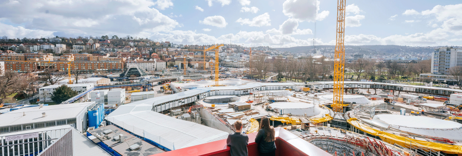 Two people stand on a roof terrace and look out over a large construction site with cranes and a city in the background., &copy; Thomas Niederm&uuml;ller