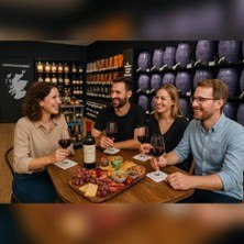 Four people sit at a table with a wine and cheese platter in a store with shelves full of bottles., &copy; see on the left