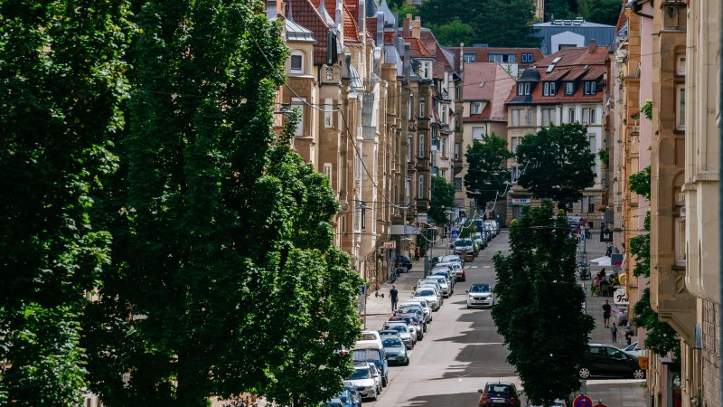 Eine belebte Stadtstraße mit geparkten Autos, alten Gebäuden und Bäumen. Im Hintergrund sind Hügel und weitere Häuser zu sehen., © Thomas Niedermüller