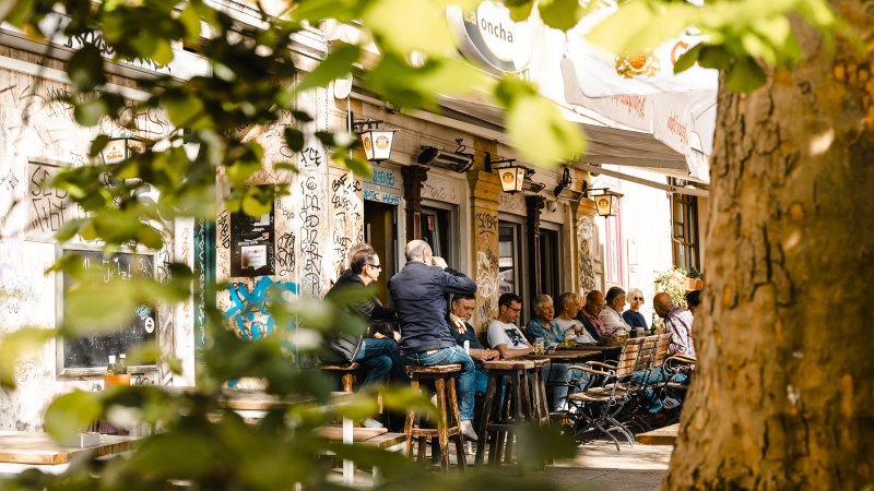 Menschen sitzen im Außenbereich eines Cafés namens La Concha, umgeben von Graffiti und Bäumen. Die Szene wirkt lebendig und entspannt., © SMG Stuttgart Marketing GmbH - Sarah Schmid