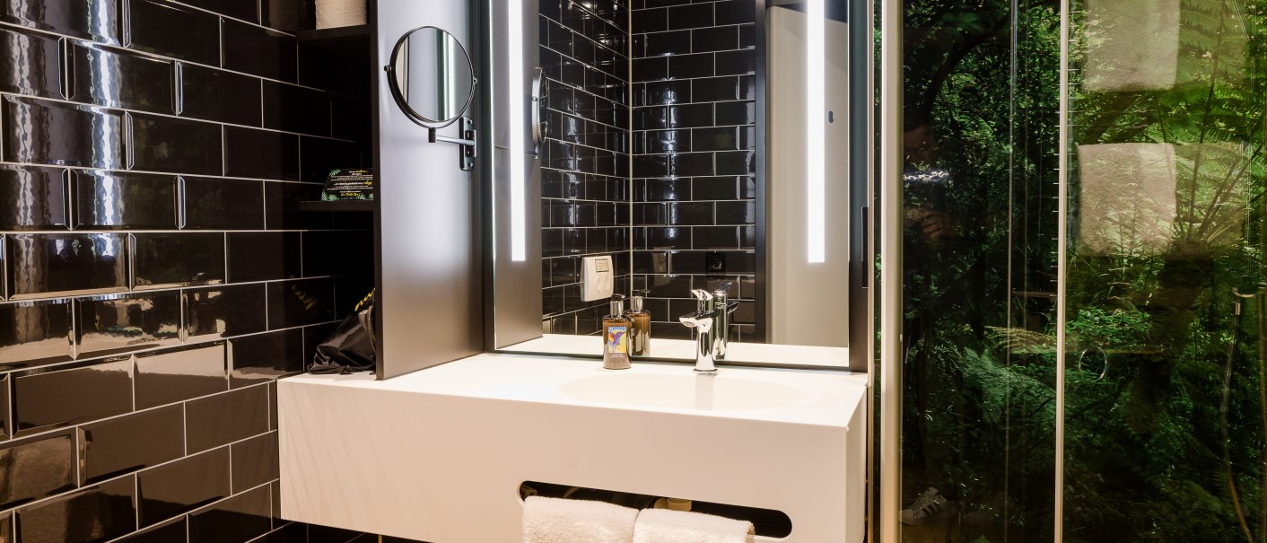 Modernes Badezimmer mit schwarzen Fliesen, beleuchtetem Spiegel und weißem Waschbecken. Ein Handtuch hängt am Waschbecken, daneben ist eine Glaswand mit Blick ins Grüne., © TOMAS