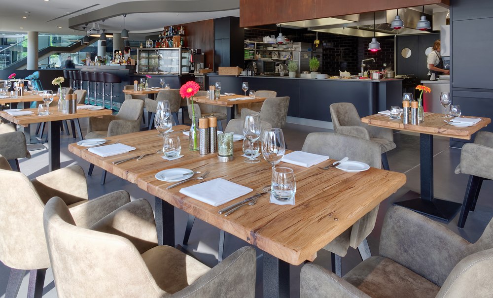 Modernes Restaurant mit Holztischen, gedeckt mit Gläsern und Besteck. Im Hintergrund eine offene Küche und Bar., © V8 HOTEL Motorworld Region Stuttgart