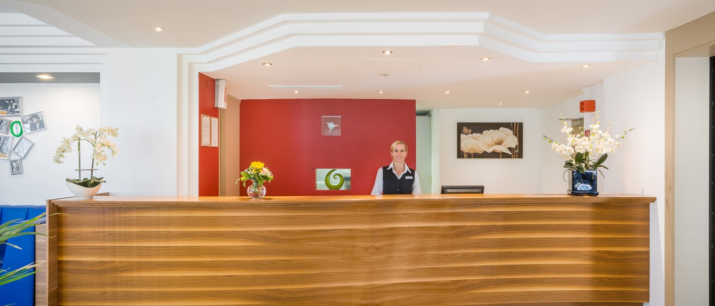 A hotel reception with a wooden reception desk. An employee stands behind it. Flowers and decorations are placed on the counter., © TOMAS