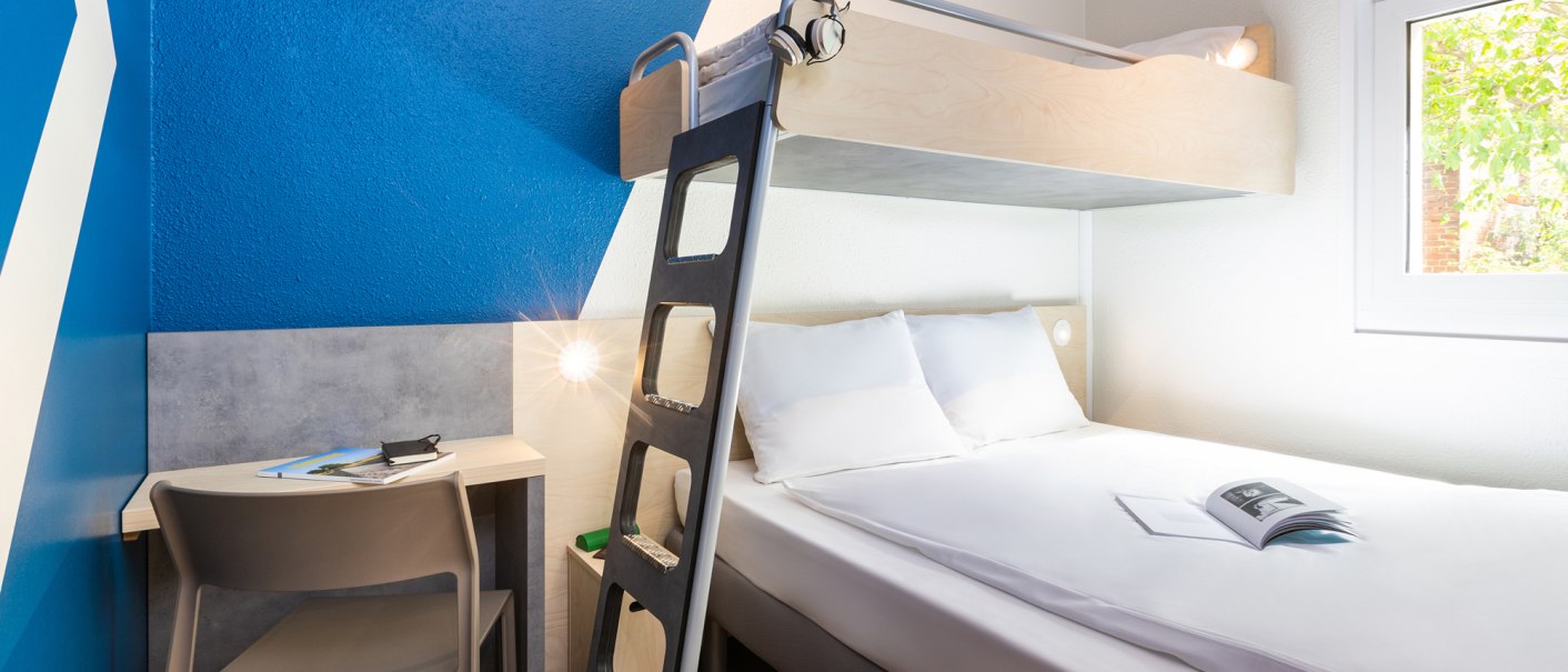 Modernes Hotelzimmer mit Doppelbett und darüberliegendem Einzelbett, blau-weiße Wand, kleiner Schreibtisch mit Stuhl, Fenster mit Blick ins Grüne., © DQuadrat Living GmbH Modernes Hotelzimmer mit Doppelbett und darüberliegendem Einzelbett, blau-weiße Wand, kleiner Schreibtisch mit Stuhl, Fenster mit Blick ins Grüne., © DQuadrat Living GmbH