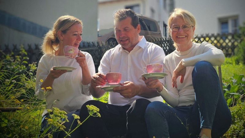 From left to right: Annette Ehrlich, Holger Köckritz and Annette Currle sit in the garden and enjoy tea from pink cups. They are wearing white tops and smiling at the camera., © Perle Uhlbach From left to right: Annette Ehrlich, Holger Köckritz and Annette Currle sit in the garden and enjoy tea from pink cups. They are wearing white tops and smiling at the camera., © Perle Uhlbach