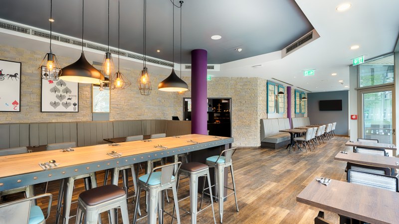 Moderner Fr&uuml;hst&uuml;cksbereich mit langen Holztischen, Metallst&uuml;hlen und stilvollen H&auml;ngelampen. Steinw&auml;nde und gro&szlig;e Fenster schaffen eine einladende Atmosph&auml;re., &copy; Premier Inn Germany