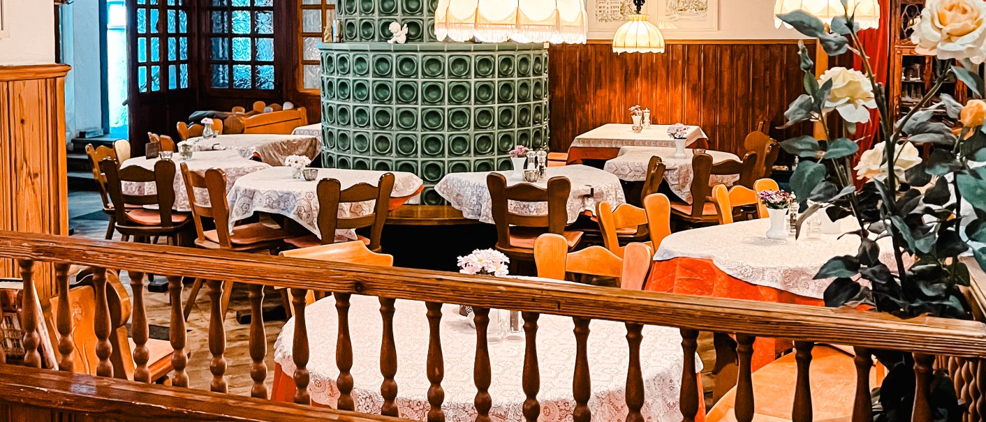 Gemütlicher Gastraum mit Holzmöbeln, einem grünen Kachelofen und dekorativen Tischdecken. Hängelampen und Blumenarrangements ergänzen das Ambiente., © Weinstube Kachelofen Gemütlicher Gastraum mit Holzmöbeln, einem grünen Kachelofen und dekorativen Tischdecken. Hängelampen und Blumenarrangements ergänzen das Ambiente., © Weinstube Kachelofen