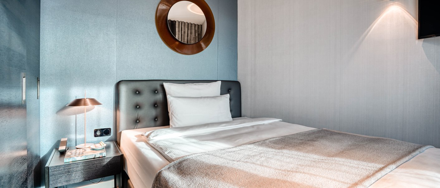 Modernes Hotelzimmer mit grauem Bett, Nachttischlampe und rundem Spiegel an der Wand. Elegantes und minimalistisches Design., © CLOUD N°7 APARTMENTS GmbH