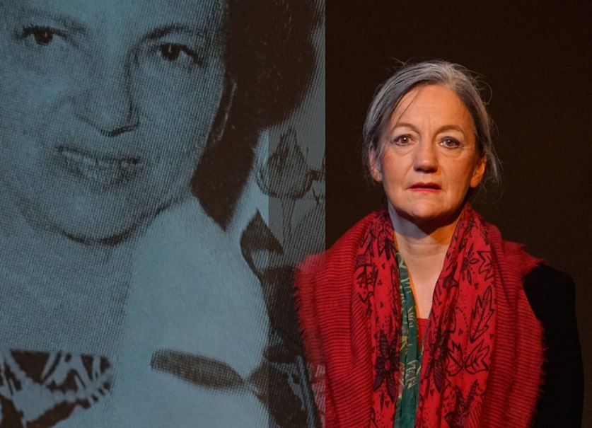 Eine Frau mit grauen Haaren und rotem Schal steht vor einem projizierten Schwarz-Weiß-Bild einer anderen Frau., © © Katja Ritter Eine Frau mit grauen Haaren und rotem Schal steht vor einem projizierten Schwarz-Weiß-Bild einer anderen Frau., © © Katja Ritter