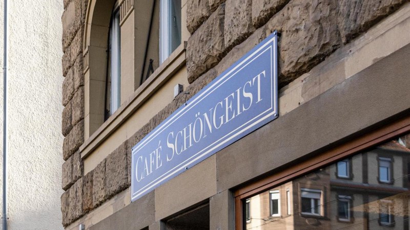 Das Bild zeigt das Schild des Caf&eacute;s Sch&ouml;ngeist an einer steinernen Hausfassade. Im Fenster spiegelt sich ein gegen&uuml;berliegendes Geb&auml;ude., &copy; Stuttgart-Marketing GmbH, Sarah Schmid