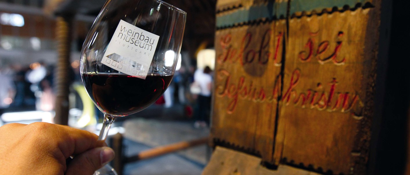 A glass of red wine with the logo of the Stuttgart Wine Museum, in the background an old wooden press with the inscription 'Praised be Jesus Christ'., &copy; Stuttgart-Marketing GmbH
