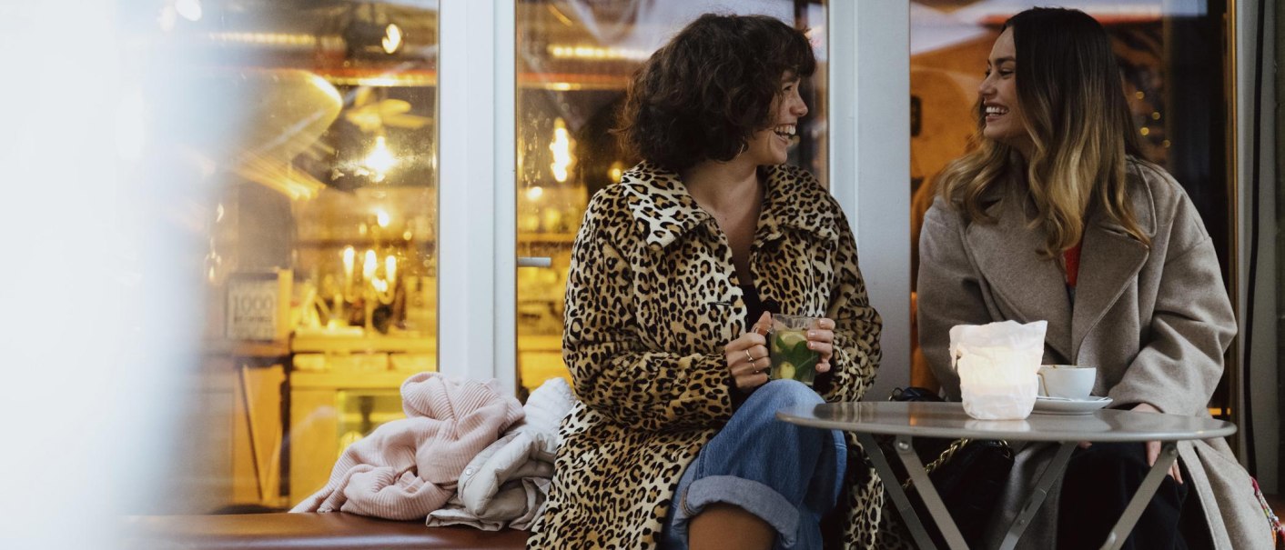 Zwei Frauen sitzen in einem Café, lachen und unterhalten sich. Eine trägt einen Leopardenmantel, die andere einen grauen Mantel. Auf dem Tisch stehen Getränke., © Stuttgart-Marketing GmbH, Alwin Maigler