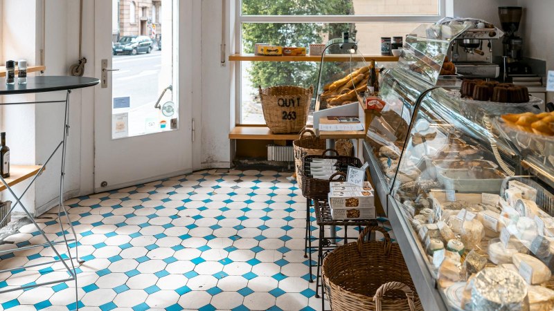 A delicatessen with cheese display, pastries and bottles of wine. Light-colored tiles and a window with a view of the street., © SMG, Sarah Schmid