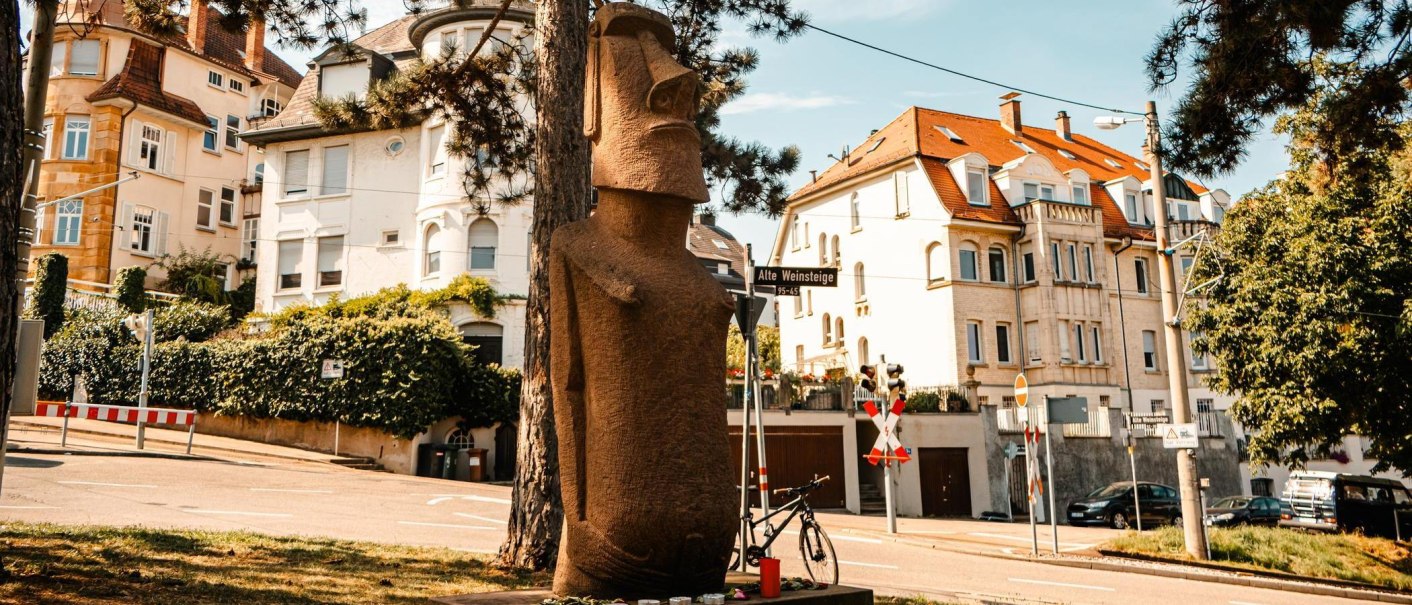 Eine Moai-Statue steht an einer Straßenecke, umgeben von Bäumen und Wohnhäusern. Ein Fahrrad lehnt an der Statue., © Stuttgart-Marketing GmbH, Sarah Schmid