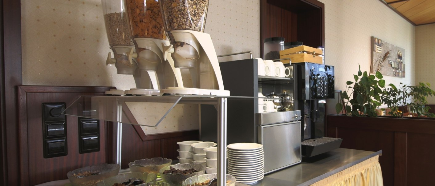 Breakfast buffet with muesli dispensers, bowls and coffee machine. Plates and cups are stacked, plants in the background., © TOMAS Breakfast buffet with muesli dispensers, bowls and coffee machine. Plates and cups are stacked, plants in the background., © TOMAS