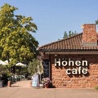 Das Höhencafé im Killesbergpark zeigt ein Gebäude aus Backstein mit Terrasse, umgeben von Bäumen und Pflanzen. Weiße Sonnenschirme sind sichtbar., © Stuttgart-Marketing GmbH, Sarah Schmid Das Höhencafé im Killesbergpark zeigt ein Gebäude aus Backstein mit Terrasse, umgeben von Bäumen und Pflanzen. Weiße Sonnenschirme sind sichtbar., © Stuttgart-Marketing GmbH, Sarah Schmid