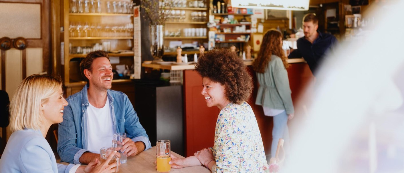 Drei Personen sitzen lachend an einem Tisch in einer gem&uuml;tlichen Bar. Im Hintergrund sind Gl&auml;ser und Flaschen zu sehen., &copy; Stuttgart-Marketing GmbH, Alwin Maigler