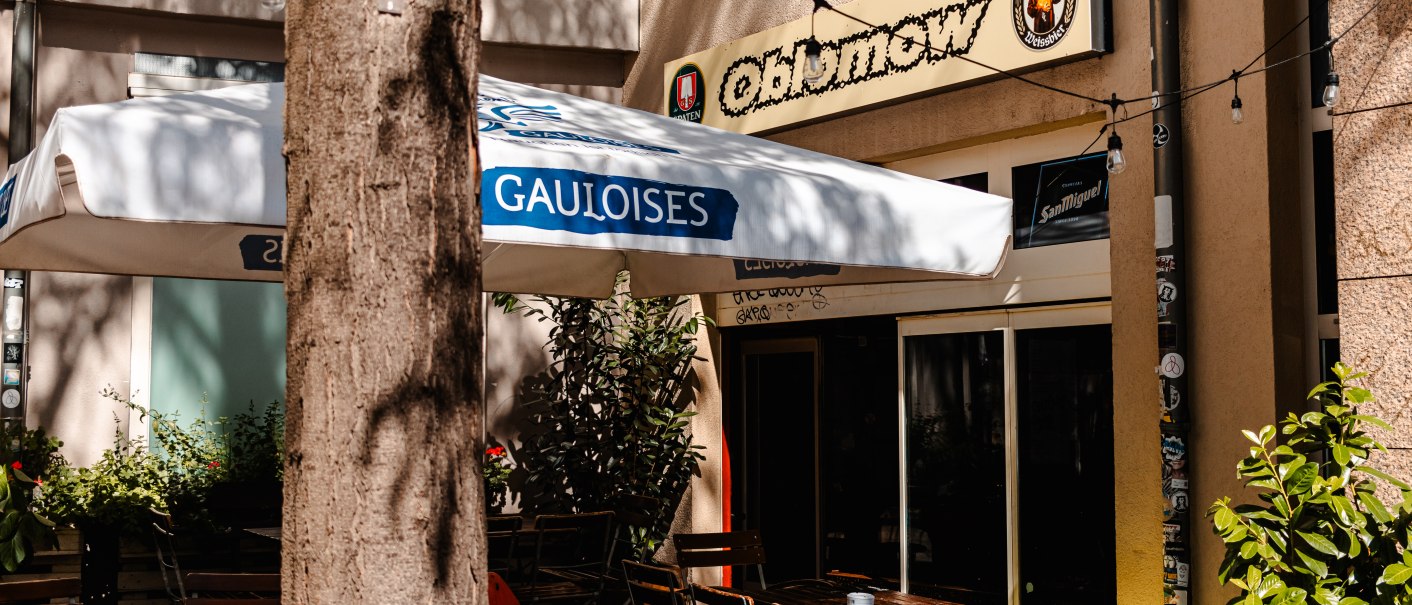 Außenbereich eines Cafés namens Oblomow mit Holztischen, Stühlen und einem Gauloises-Sonnenschirm. Ein Baum steht im Vordergrund., © SMG_Sarah Schmid