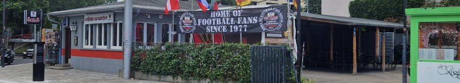 Bar mit Fahnen und Schild "Home of Football Fans Since 1977". Gebäude mit roten und grauen Akzenten, umgeben von Bäumen und einem Parkplatz., © Stuttgart-Marketing GmbH