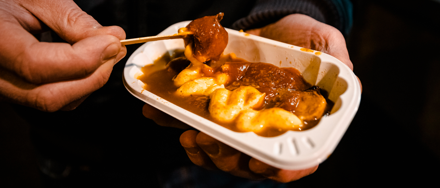 Nahaufnahme von Händen, die eine Schale mit Currywurst und Pommes halten. Ein Holzspieß wird verwendet, um ein Stück Wurst aufzuspießen., © Stuttgart-Marketing GmbH, Sarah Schmid