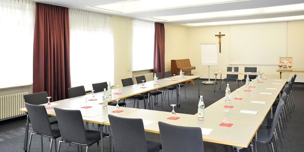 Konferenzraum mit U-förmiger Tischanordnung, Stühlen, Wasserflaschen und Gläsern. Ein Kreuz an der Wand, Flipchart und Klavier im Hintergrund., © Christkönigshaus