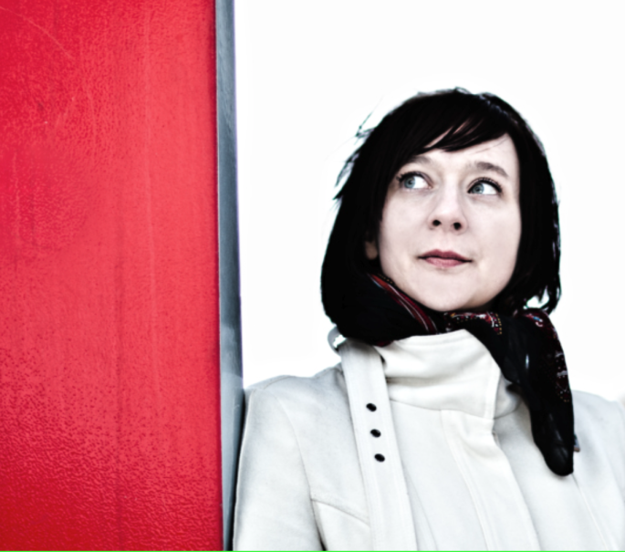 Eine Frau mit dunklem Haar und hellem Mantel steht neben einer roten Wand und blickt nach rechts. Der Hintergrund ist wei&szlig; und minimalistisch., &copy; privat