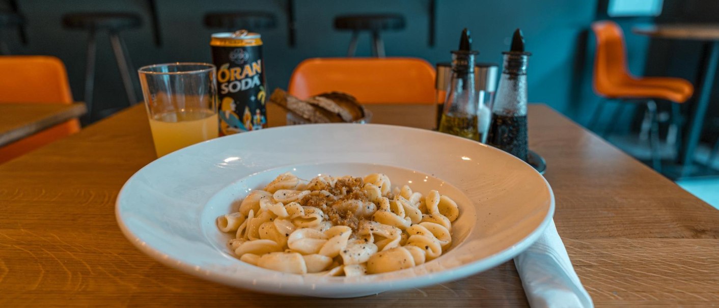 Ein Teller Pasta mit Soße, ein Glas Orangensaft und eine Dose Oran Soda auf einem Tisch in einem Restaurant., © SMG, Sarah Schmid