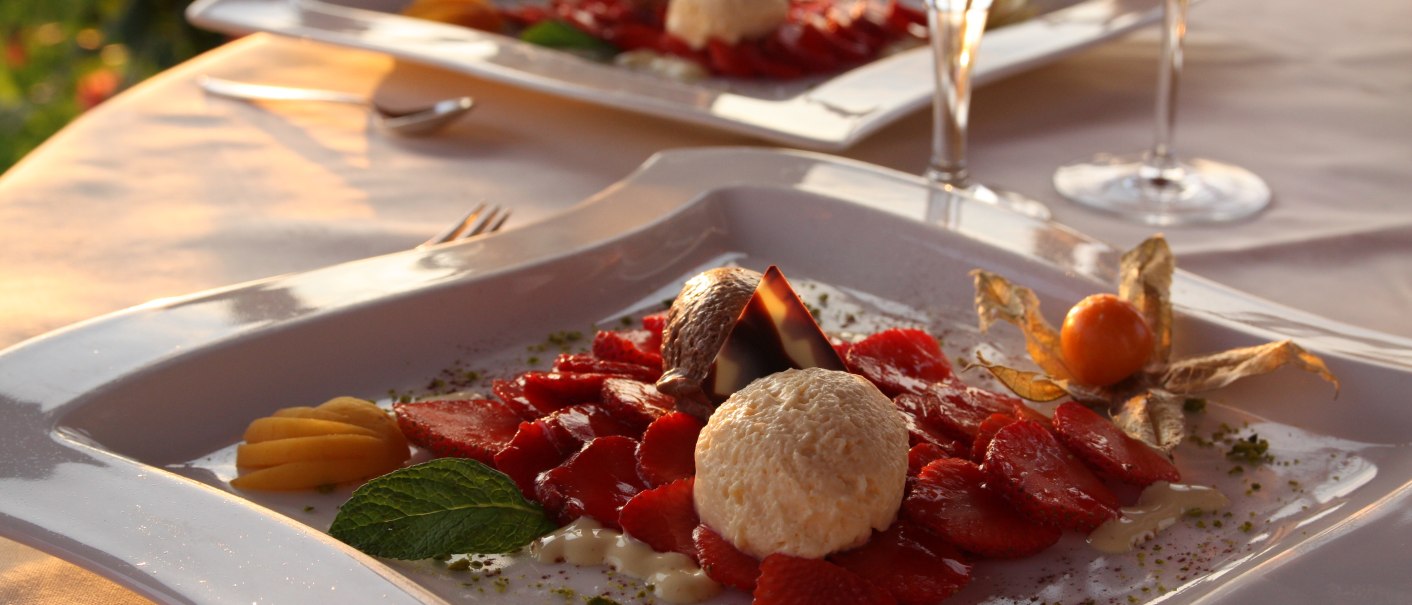 Elegantes Dessert mit Erdbeeren, Sahne und Schokolade, dekoriert mit Physalis und Minze, auf einem stilvollen Teller serviert. Im Hintergrund Weingläser., © Badhotel Restaurant Stauferland