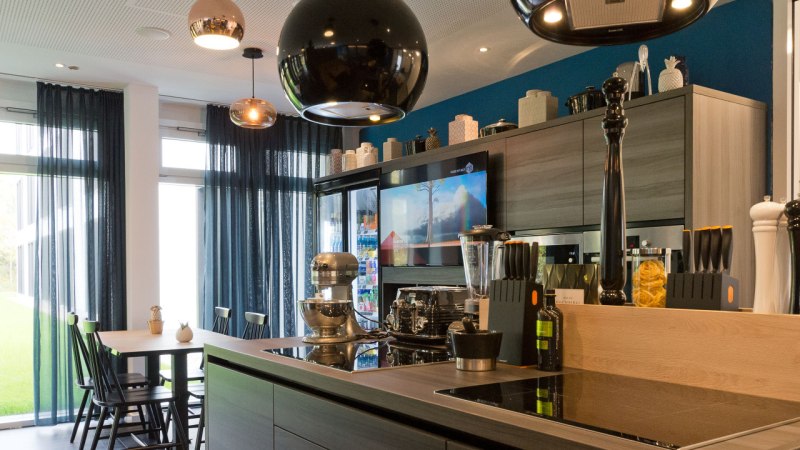 Moderne Küche mit Kochinsel, Esstisch, Hängelampen und Geräten. Große Fenster mit blauen Vorhängen lassen viel Licht herein., © Arthotel Ana / elaya Moderne Küche mit Kochinsel, Esstisch, Hängelampen und Geräten. Große Fenster mit blauen Vorhängen lassen viel Licht herein., © Arthotel Ana / elaya