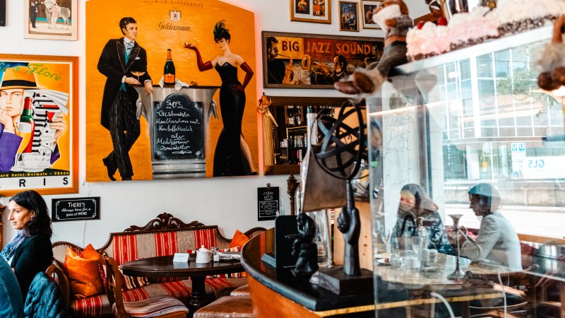 Ein gemütliches Café mit bunten Postern, darunter ein großes Bild mit einem Paar und Champagner. Menschen sitzen an Tischen, umgeben von Dekorationen., © SMG_Sarah Schmid Ein gemütliches Café mit bunten Postern, darunter ein großes Bild mit einem Paar und Champagner. Menschen sitzen an Tischen, umgeben von Dekorationen., © SMG_Sarah Schmid