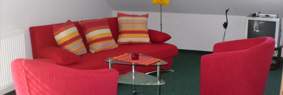 Ein Wohnzimmer mit roten Sesseln und Sofa, gestreiften Kissen, Glastisch, grünem Teppich und Fernseher in einer Dachgeschosswohnung., © Weinhof am Württemberg Ein Wohnzimmer mit roten Sesseln und Sofa, gestreiften Kissen, Glastisch, grünem Teppich und Fernseher in einer Dachgeschosswohnung., © Weinhof am Württemberg