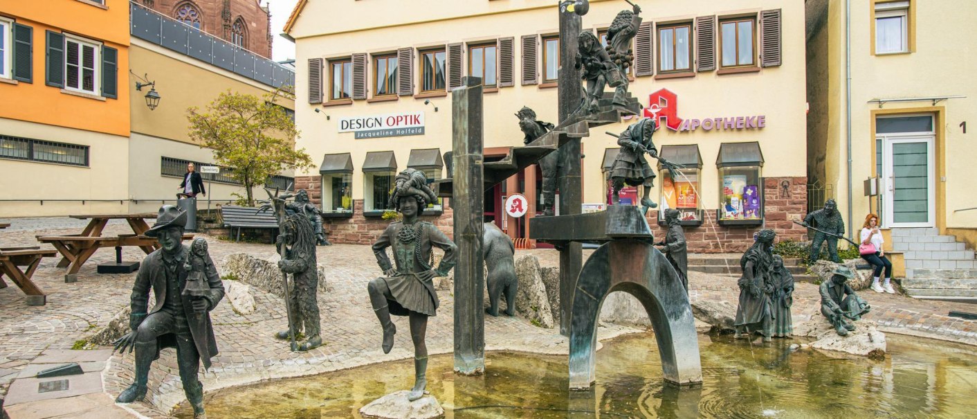 The Fools' Fountain in Weil der Stadt shows bronze figures of fools in various poses. Stores and a pharmacy can be seen in the background., © Stuttgart-Marketing GmbH, Sarah Schmid