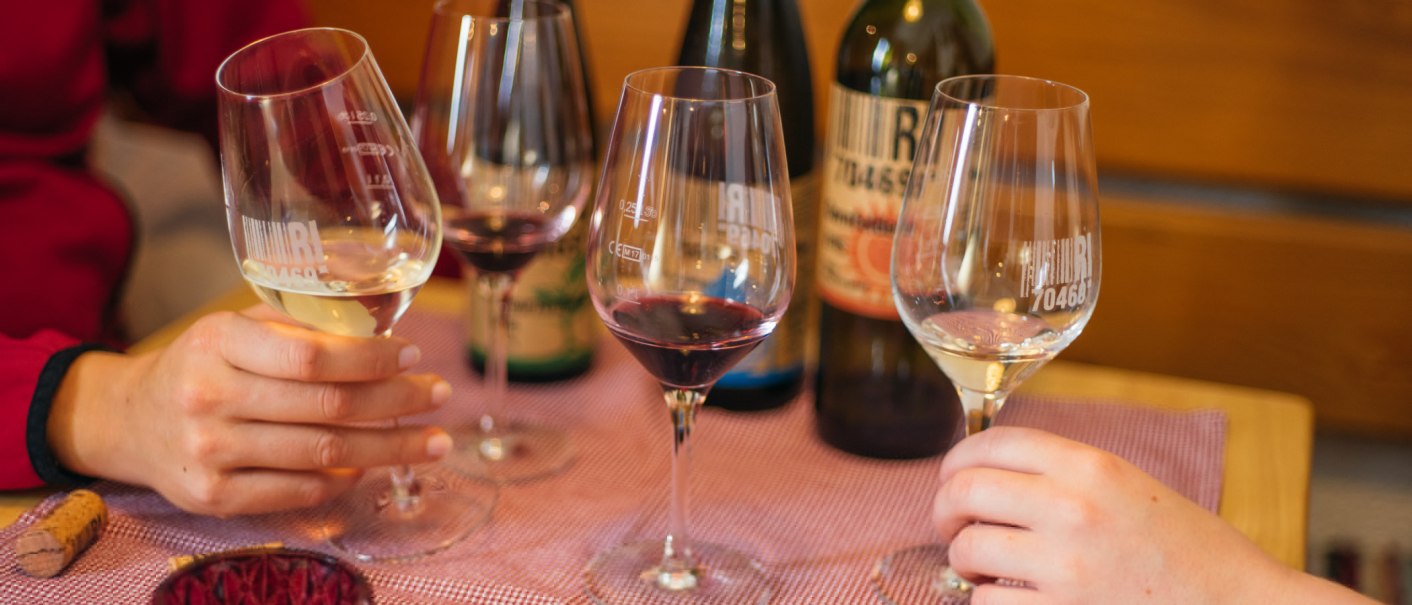 Two people are holding glasses of red and white wine, clinking glasses. In the background are bottles of wine on a table with a purple tablecloth., © 70469R! Weinbau Fabian Rajtschan Two people are holding glasses of red and white wine, clinking glasses. In the background are bottles of wine on a table with a purple tablecloth., © 70469R! Weinbau Fabian Rajtschan