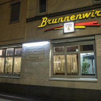 Das Restaurant Brunnenwirt bei Nacht, beleuchtet mit gelbem Schriftzug. Ein Mann steht an einem Tisch vor dem Eingang., © SMG, Sarah Schmid Das Restaurant Brunnenwirt bei Nacht, beleuchtet mit gelbem Schriftzug. Ein Mann steht an einem Tisch vor dem Eingang., © SMG, Sarah Schmid