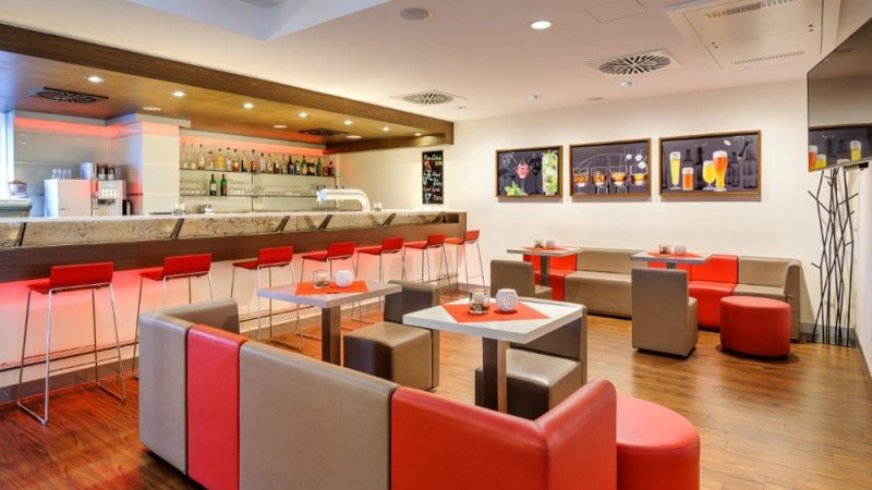 Moderne Bar mit roten und grauen Möbeln, beleuchtetem Tresen und Wandbildern von Getränken. Elegantes Ambiente mit Holzboden., © Ibis Stuttgart Airport Messe