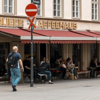 Ein Café mit rotem Vordach und dem Schriftzug 'Queer Kaffeehaus'. Menschen sitzen draußen an Tischen. Ein Mann geht vorbei., © SMG, Sarah Schmid Ein Café mit rotem Vordach und dem Schriftzug 'Queer Kaffeehaus'. Menschen sitzen draußen an Tischen. Ein Mann geht vorbei., © SMG, Sarah Schmid