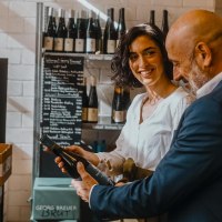 Two people in a wine shop, a woman smiling while holding a bottle of wine. Wines and a price board can be seen in the background., © SMG, Alwin Maigler Two people in a wine shop, a woman smiling while holding a bottle of wine. Wines and a price board can be seen in the background., © SMG, Alwin Maigler