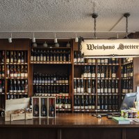 Counter at Weinhaus Stetter with shelves full of wine bottles and a wooden counter. A sign reading 'Weinhaus Stetter' hangs from the ceiling., © SMG Stuttgart Marketing GmbH - Sarah Schmid Counter at Weinhaus Stetter with shelves full of wine bottles and a wooden counter. A sign reading 'Weinhaus Stetter' hangs from the ceiling., © SMG Stuttgart Marketing GmbH - Sarah Schmid