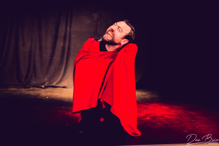 Ein Schauspieler in rotem Umhang kniet auf der Bühne und zeigt eine leidenschaftliche Geste. Der Hintergrund ist dunkel gehalten., © Theater am Olgaeck/Dan Boia Ein Schauspieler in rotem Umhang kniet auf der Bühne und zeigt eine leidenschaftliche Geste. Der Hintergrund ist dunkel gehalten., © Theater am Olgaeck/Dan Boia