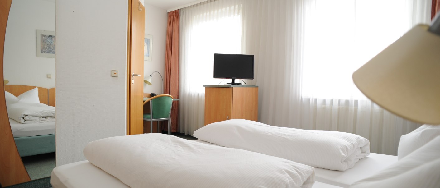 A hotel room with two single beds, a TV on a wardrobe, a desk with chair and large windows with curtains., © TOMAS