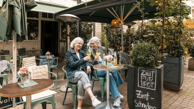 Zwei Frauen sitzen in einem gem&uuml;tlichen Caf&eacute; im Freien, sto&szlig;en mit Getr&auml;nken an und lachen. Ein Schild bietet Zimtschnecken an., &copy; Stuttgart Marketing GmbH - Sarah Schmid