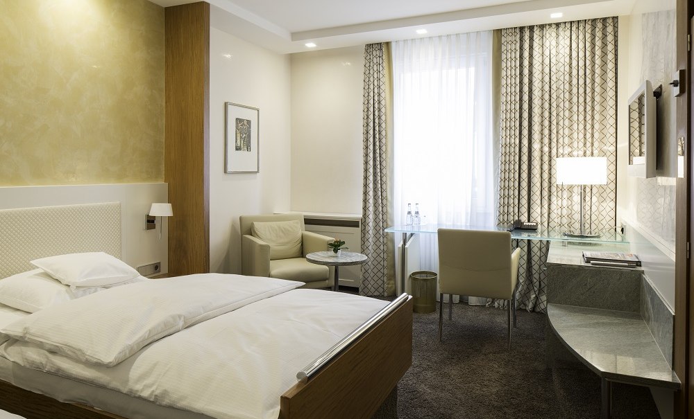 Modernes Einzelzimmer mit Bett, Schreibtisch, Sessel und Fenster mit Vorhängen. Helle Farben und stilvolle Einrichtung schaffen eine gemütliche Atmosphäre., © Hotel Royal