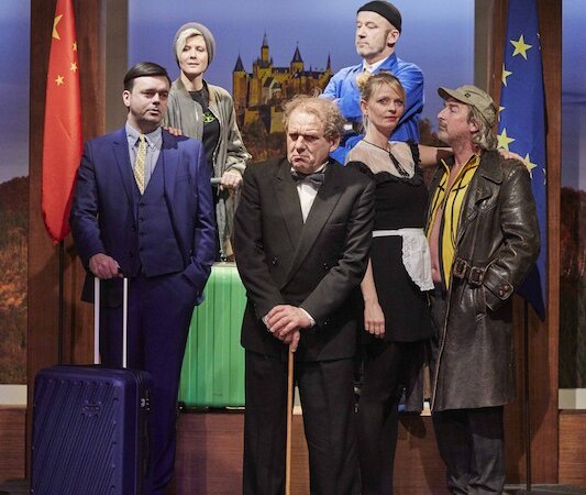Theaterszene mit sechs Personen in Kostümen vor Flaggen und einem Schlossbild. Ein Mann im Anzug hält einen Koffer, eine Frau trägt ein Dienstmädchen-Outfit., © Wilhelma Theater Theaterszene mit sechs Personen in Kostümen vor Flaggen und einem Schlossbild. Ein Mann im Anzug hält einen Koffer, eine Frau trägt ein Dienstmädchen-Outfit., © Wilhelma Theater