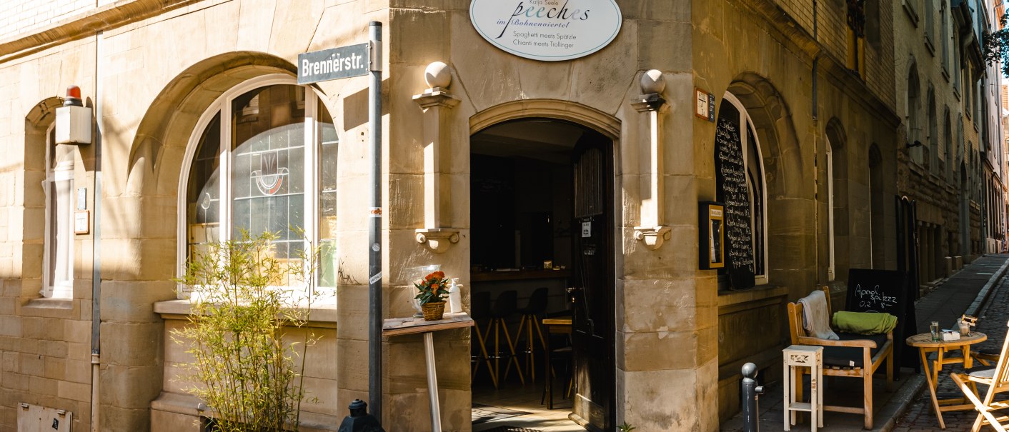Ein gemütliches Außencafé an einer Straßenecke mit Holzmöbeln, Pflanzen und einem Straßenschild. Die Sonne scheint auf die Fassade., © SMG Stuttgart Marketing GmbH - Sarah Schmid