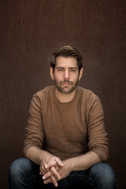 A man in a brown sweater sits with his hands clasped in front of a brown background and looks seriously into the camera., &copy; Renitenztheater Stuttgart e.V.