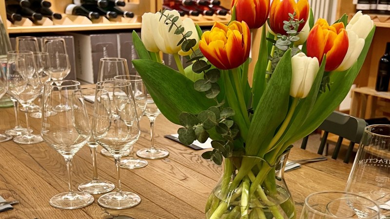 Ein Tisch mit Weingläsern und einem Tulpenstrauß, umgeben von Weinflaschen in Regalen. Elegante Atmosphäre für einen Weinabend., © Weinfactum Bad Cannstatt