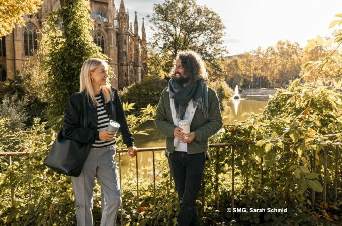 Zwei Personen spazieren l&auml;chelnd mit Kaffeebechern vor einer Kirche in Stuttgart. Im Hintergrund ein Teich mit Springbrunnen und herbstlicher Vegetation., &copy; Stuttgart Marketing GmbH