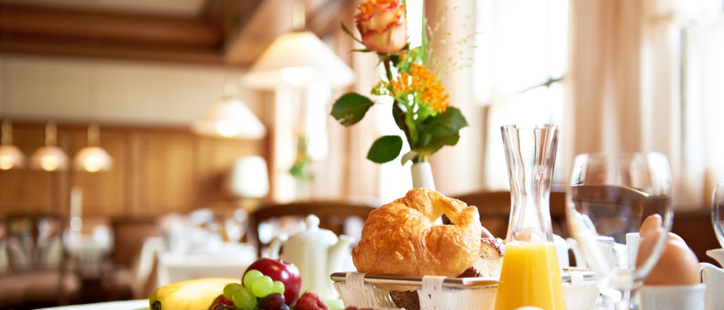 Ein Frühstückstisch mit Croissant, Obst, Orangensaft und Blumen in einem hellen Raum., © Hirsch Hotel